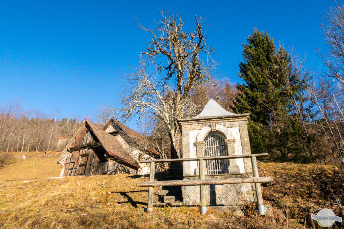 Kleines Marterl, dahinter eine Hütte und ein Baum